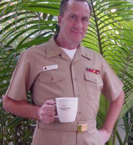 David W Bynon in a CPO khaki uniform holding a white Starbucks mug, standing in front of large green palm leaves.